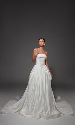 A woman stands against a dark gray background wearing the Long Wedding Dress: Alyce 7187 in Diamond White, a strapless satin gown with a full skirt, train, and classic ballgown silhouette.