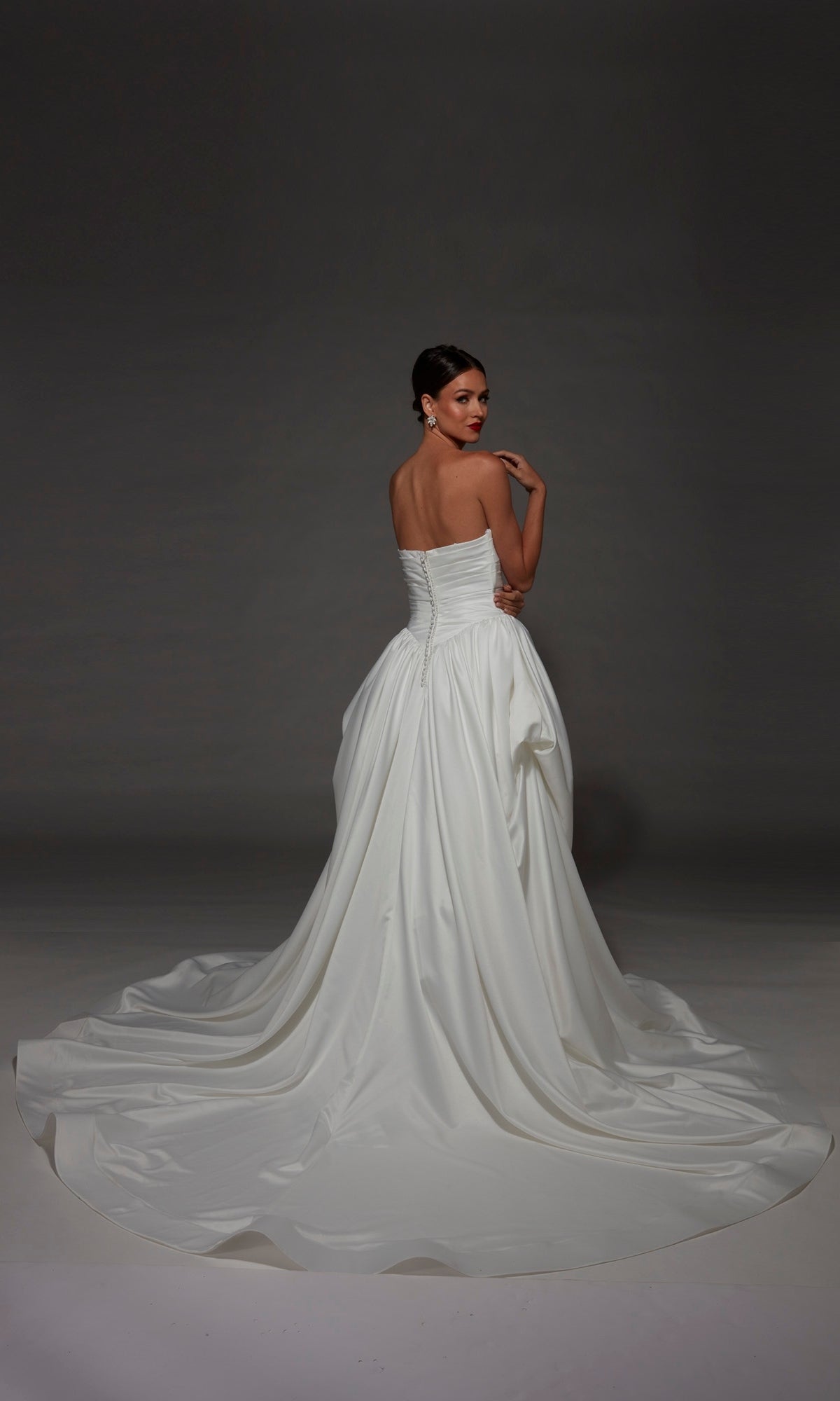 A woman wearing the Alyce 7187 long wedding dress, a strapless satin gown with a sweeping train, stands facing away and glances back over her shoulder against a dark background.
