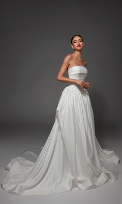 A woman wearing the Alyce 7187 long wedding dress—a strapless satin gown with a full skirt and long train—stands against a plain grey background.