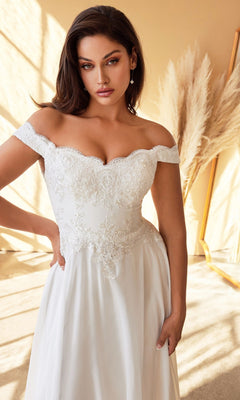 A woman stands indoors in warm lighting among neutral pampas grass, wearing the elegant and timeless Long Off-White Wedding Dress: CL7258W adorned with white lace—an affordable off-the-shoulder bridal gown.