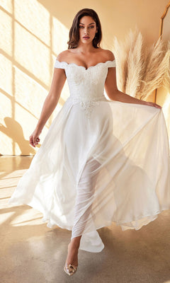 A woman wearing the CL7258W long off-white wedding dress with an off-the-shoulder lace bodice and flowing skirt stands indoors near pampas grass. This elegant, timeless gown is perfect for any bride.