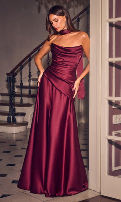 A woman stands indoors by a staircase wearing the Ladivine 7432 long prom dress in burgundy, featuring a floor-length satin gown with a ruched bodice, asymmetrical neckline, matching choker, and an elegant lace-up back for added detail and sophistication.