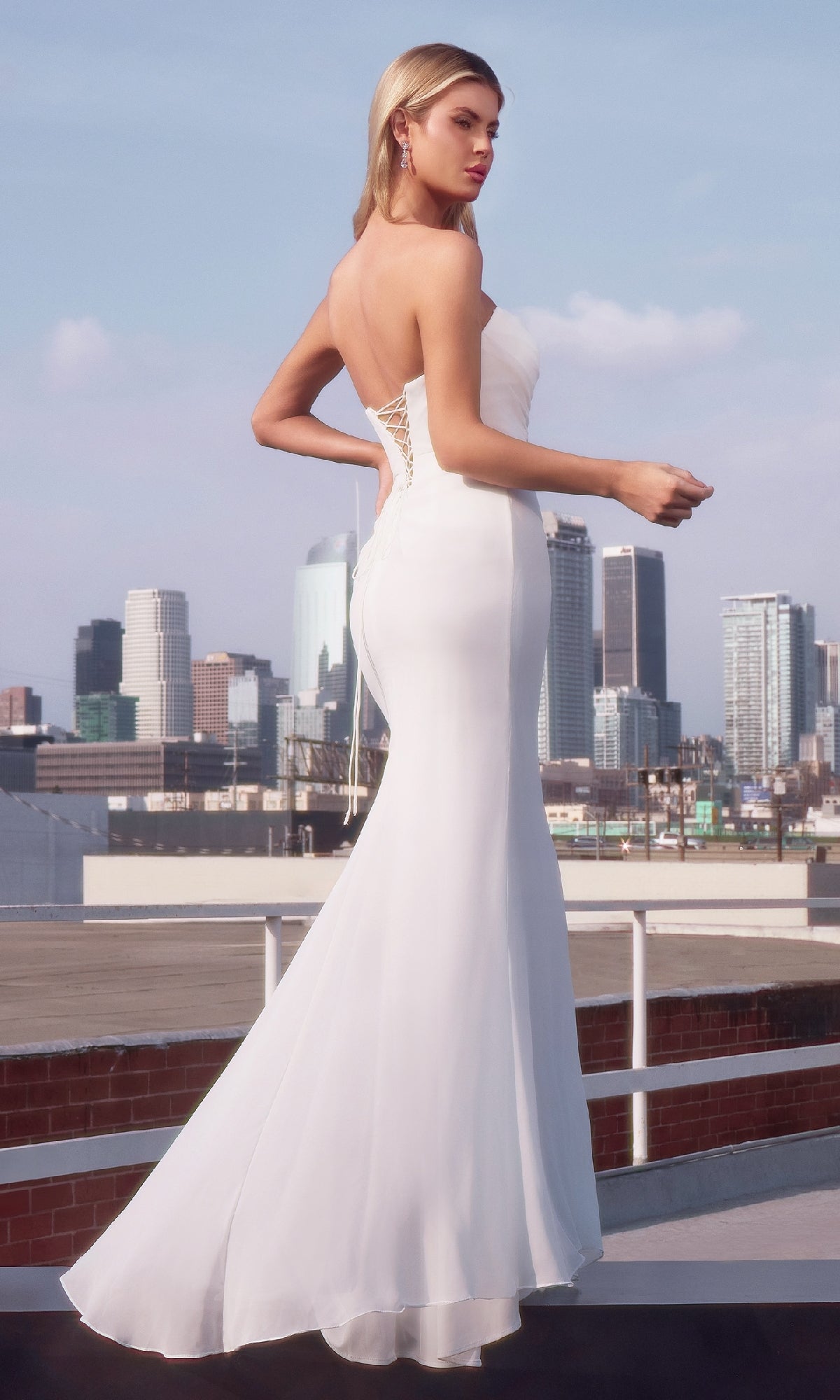 A woman in the affordable Ladivine 7434W—a strapless, form-fitting long white prom dress—stands on a rooftop with the city skyline behind her.