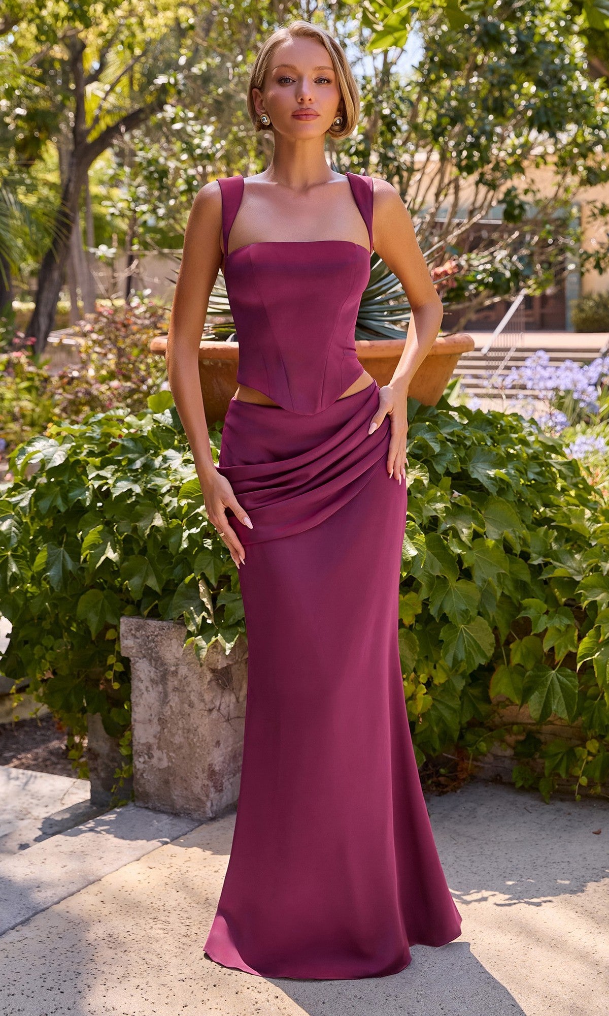 A woman stands outdoors amidst greenery and sunlight, wearing the Ladivine 7437 Long Prom Dress in mulberry—a sleeveless evening gown featuring a corset bodice, cut-out waist detail, and a draped skirt.