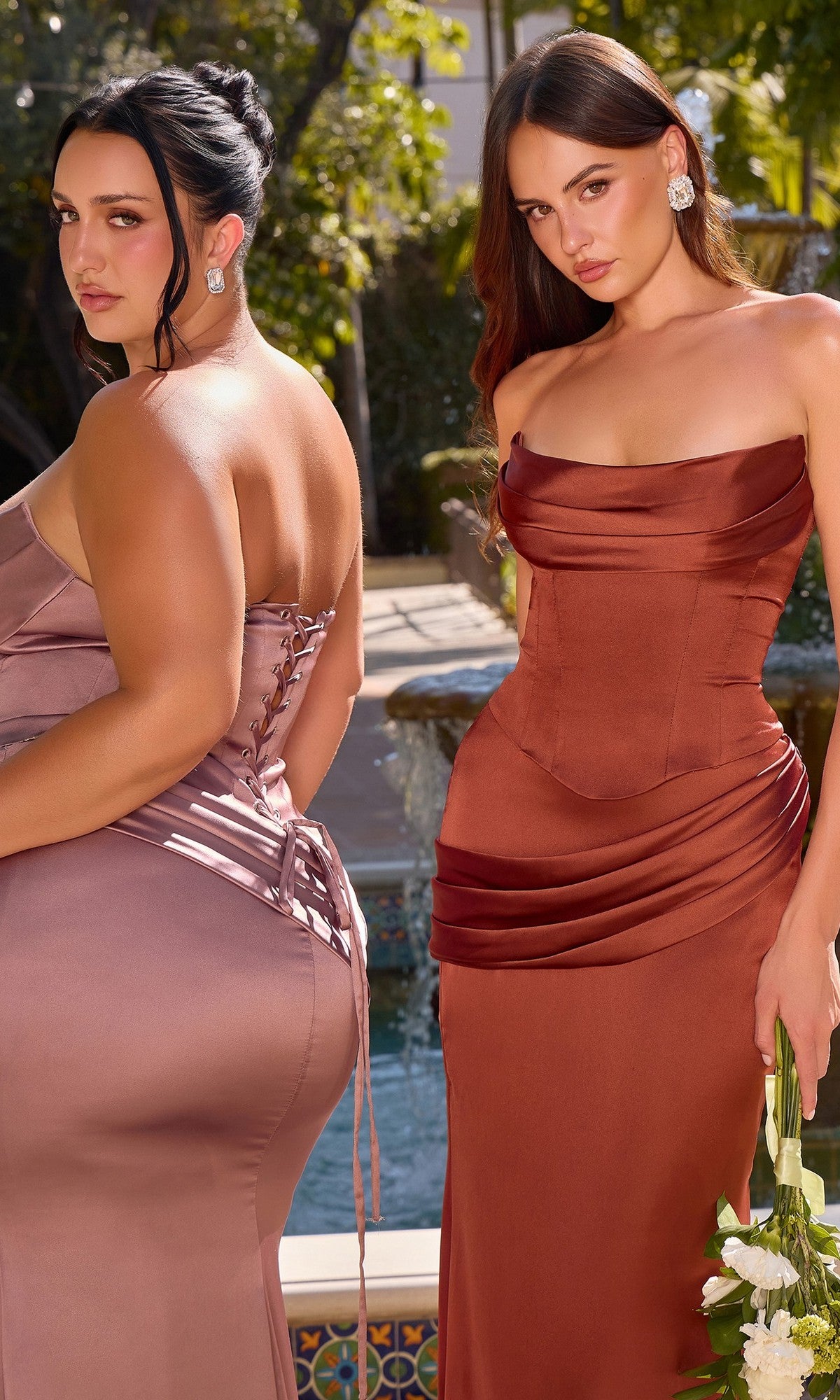 Wearing the Long Prom Dress: Ladivine 7439, two women pose outdoors—one in a mauve version with a lace-up back and contemporary silhouette, the other in a rust-colored style with draped detailing. Lush trees and an ornate fountain complete the elegant setting.