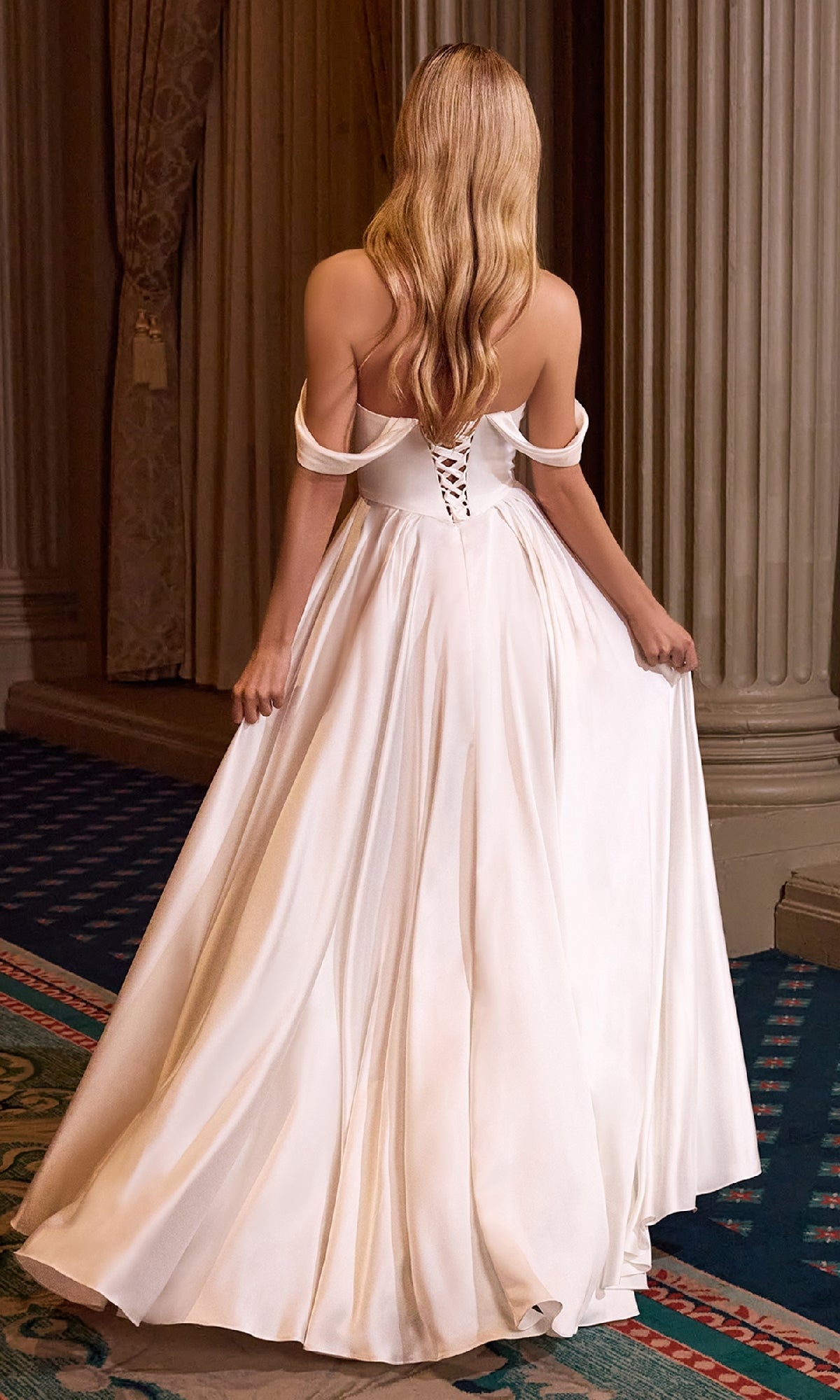 A woman with long blonde hair wears the Off-Shoulder Long White Prom Dress: Ladivine 7449W, featuring a lace-up back, standing elegantly in a room with columns and draped curtains—the perfect special occasion look.