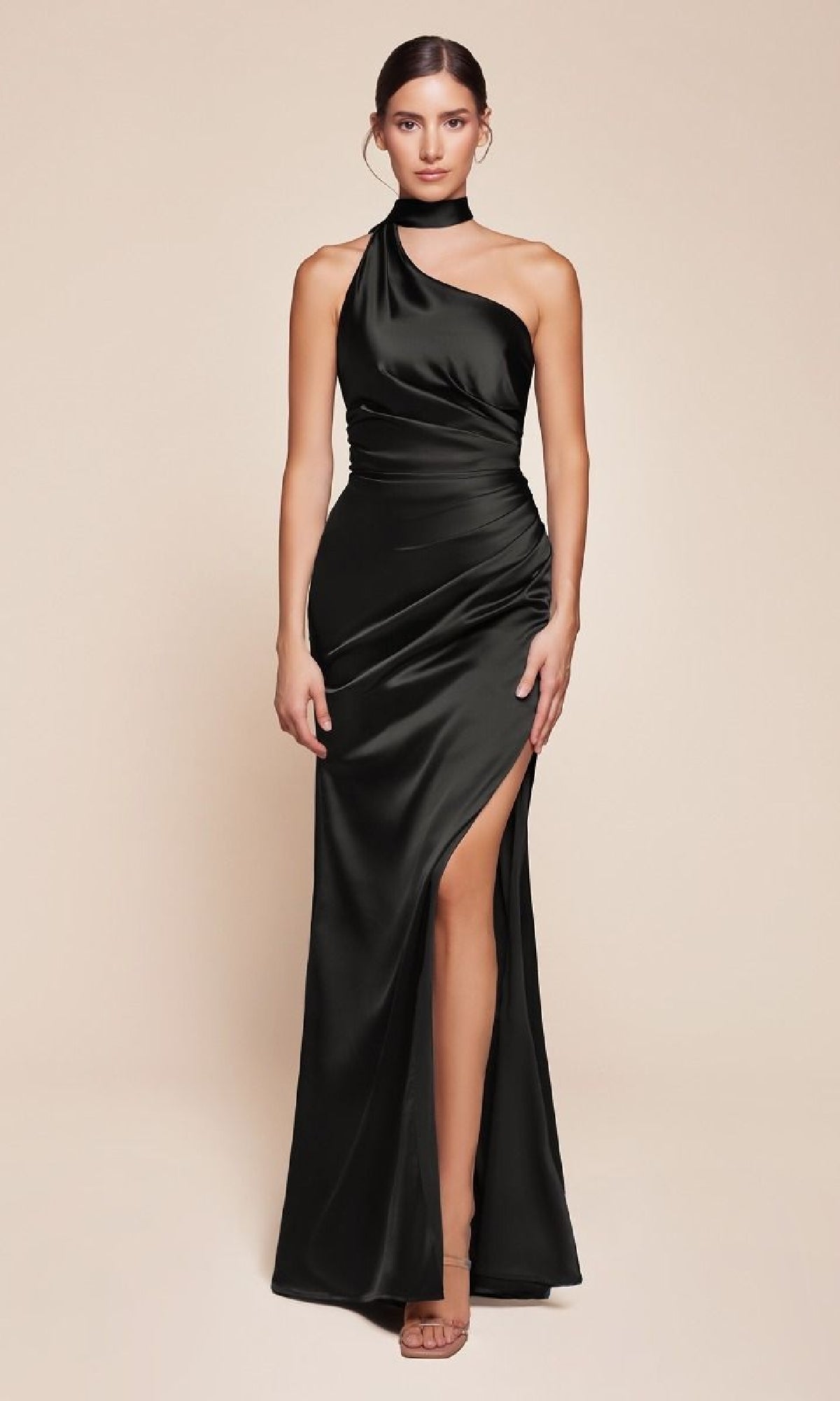 A woman poses against a beige background, wearing the Ladivine 7454 One-Shoulder Sash Long Prom Dress in black satin with a high slit.
