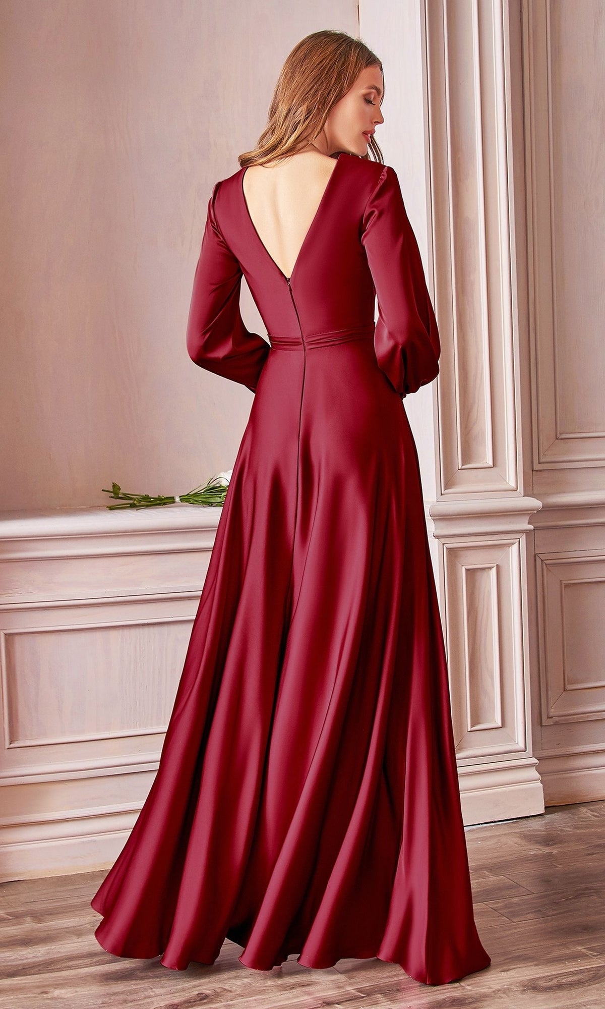 A woman stands indoors wearing the Split Long Sleeve Long Formal Dress 7475 in burgundy, its open back and flowing fabric cascading elegantly as she faces away from the camera.