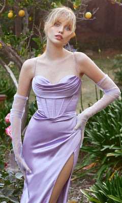 Wearing the Ladivine 7483 Long Satin Prom Dress with a corset bodice and thigh-high slit in lavender, a woman stands outdoors among greenery and flowers, her look finished with sheer gloves.