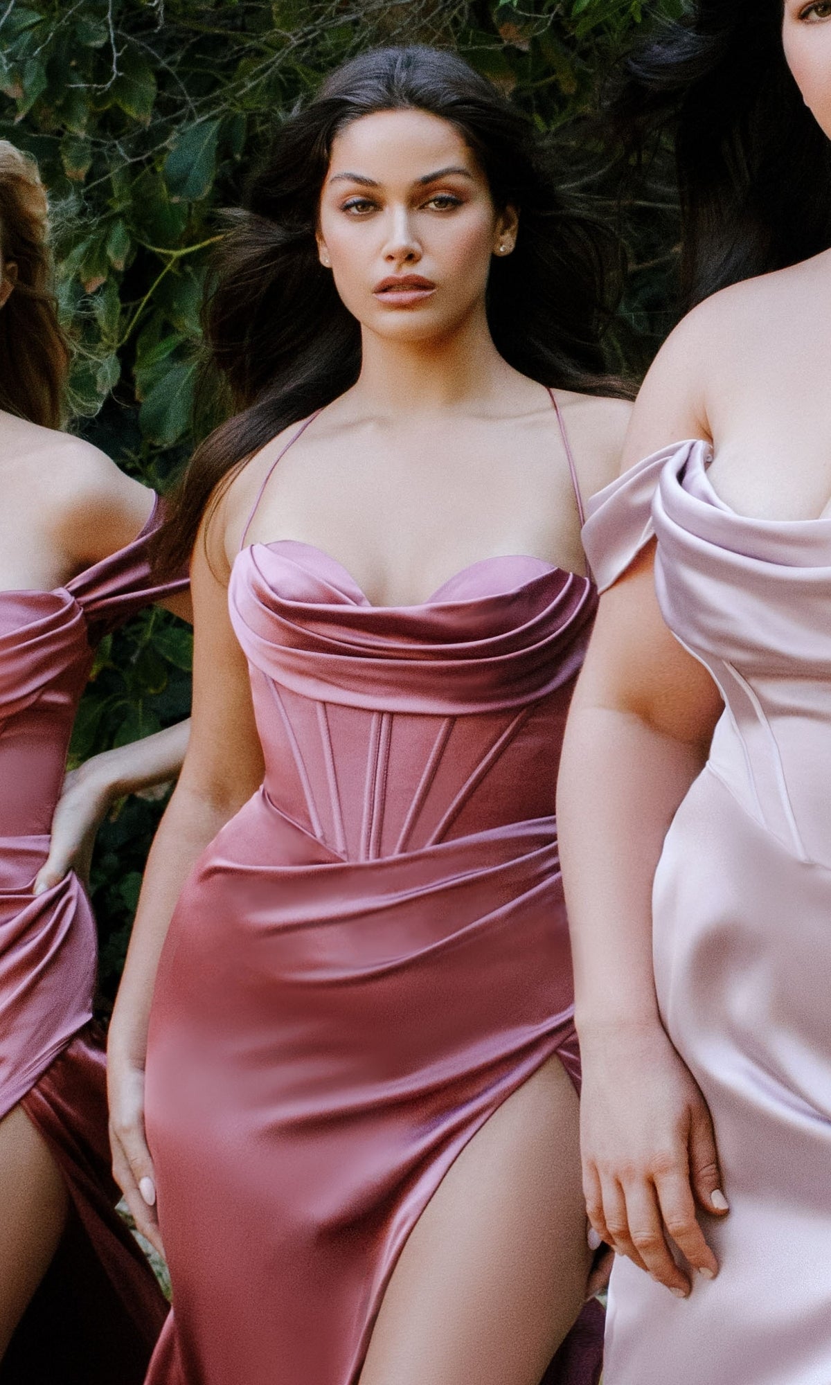 A woman outdoors wears the Ladivine 7483, a fitted pink long satin prom dress with a corset-style bodice and thigh-high slit, standing among others in similar dresses.