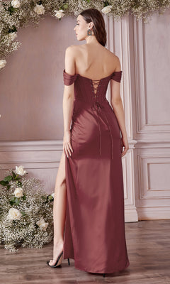 A woman stands on a wooden floor in the Off-the-Shoulder Long Plus-Size Prom Dress 7484C—a mauve gown with lace-up back and thigh-high slit—surrounded by white floral arrangements against a paneled wall.