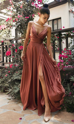 A woman in the Plus-Size Long A-Line Prom Dress 7485C, a sleeveless copper satin gown with a thigh-high slit, stands outdoors by pink flowers and greenery holding the sides of her dress.