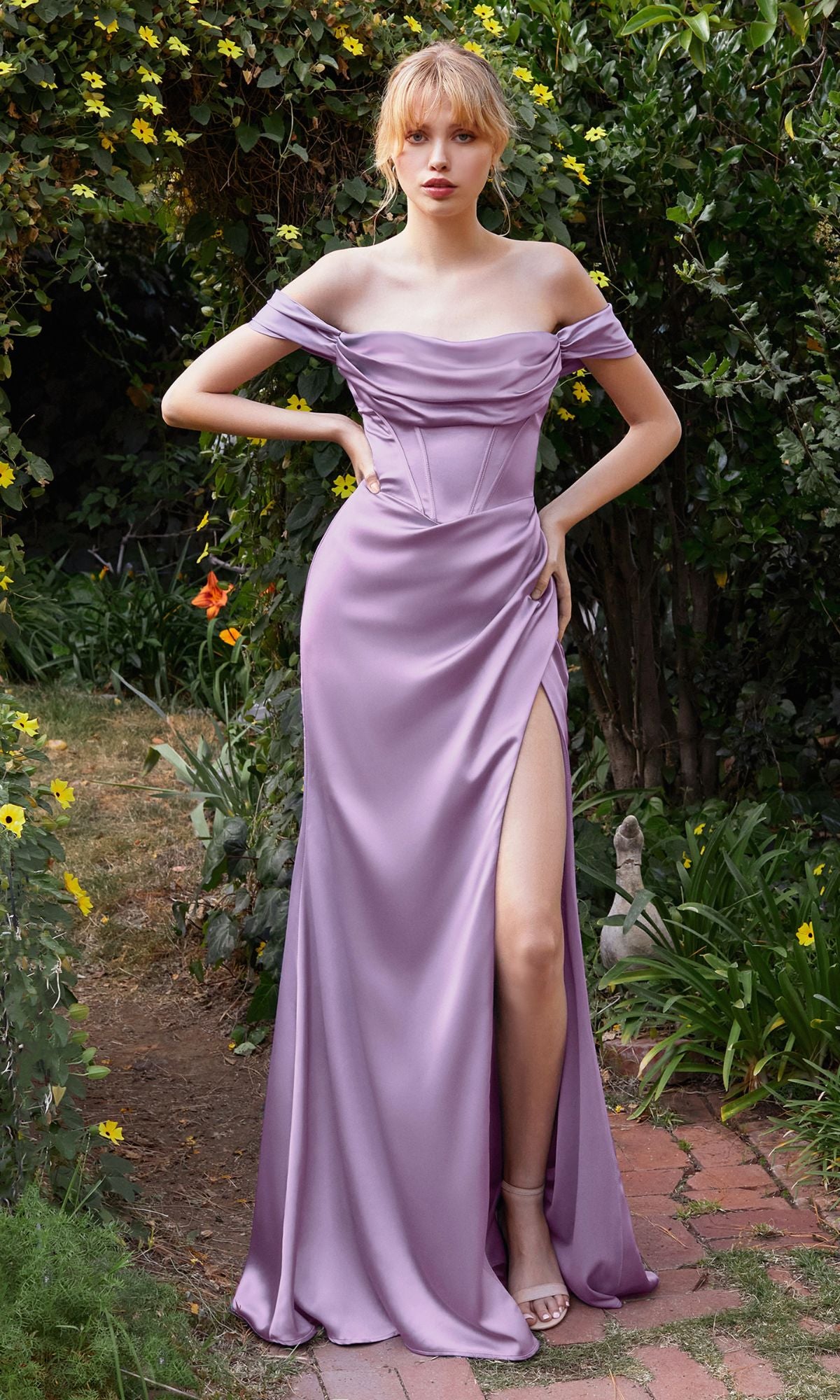 A woman stands in a garden wearing the Off-the-Shoulder Satin Prom Dress 7492 in light purple, featuring a thigh-high slit and strappy heels.