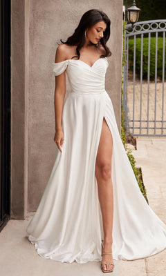 A woman poses outdoors by a stone wall and metal gate in the Ladivine 7493W, an off-shoulder white prom gown with a thigh-high slit, paired with open-toe heels.