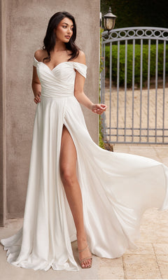 A woman wearing the Ladivine 7493W Off-Shoulder White Prom Gown, featuring a long flowing skirt and high slit, stands near a wall and iron gate, elegantly holding part of her dress.