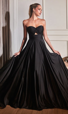 A woman stands indoors wearing the Strapless Sweetheart Long A-Line Prom Dress 7496 in black, a floor-length gown with a cutout under the bust, holding the sides of her dress and looking to the left.