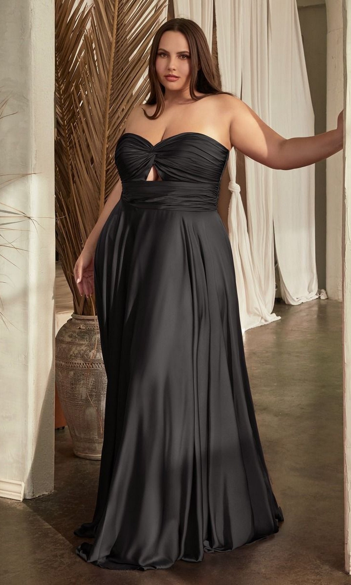 A woman wearing the Plus-Size Strapless Sweetheart Prom Dress 7496C in black stands indoors near a doorway, with draped curtains and a large decorative vase in the background.