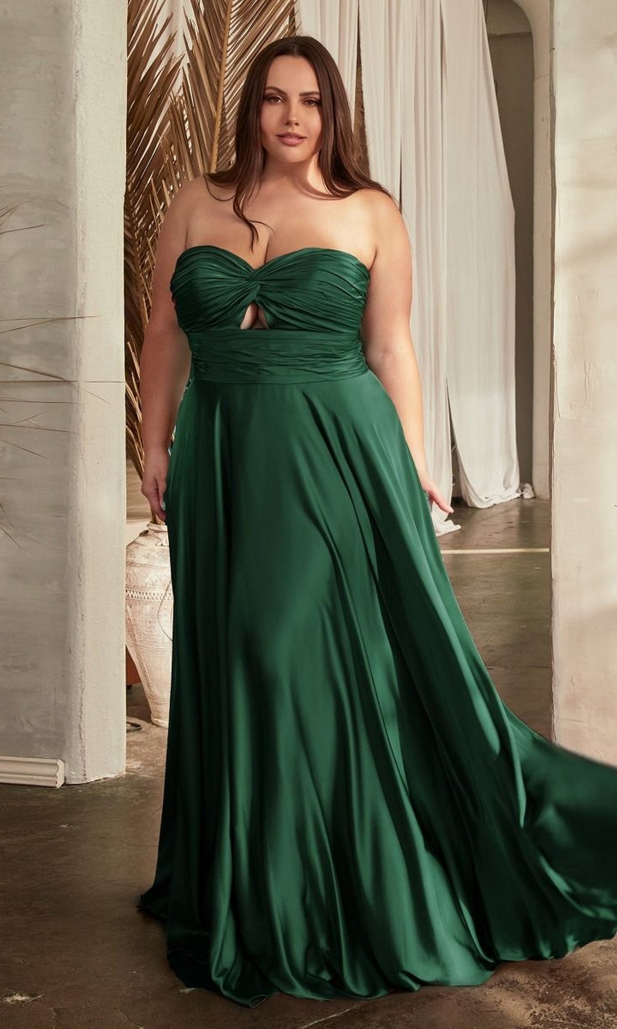A woman stands indoors near white curtains and a plant, wearing the Plus-Size Strapless Sweetheart Prom Dress 7496C in dark green.