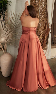 A woman models the Plus-Size Strapless Sweetheart Prom Dress 7496C in rust satin, standing with her back to the camera and arms raised, surrounded by dried palm leaves and draped fabric.