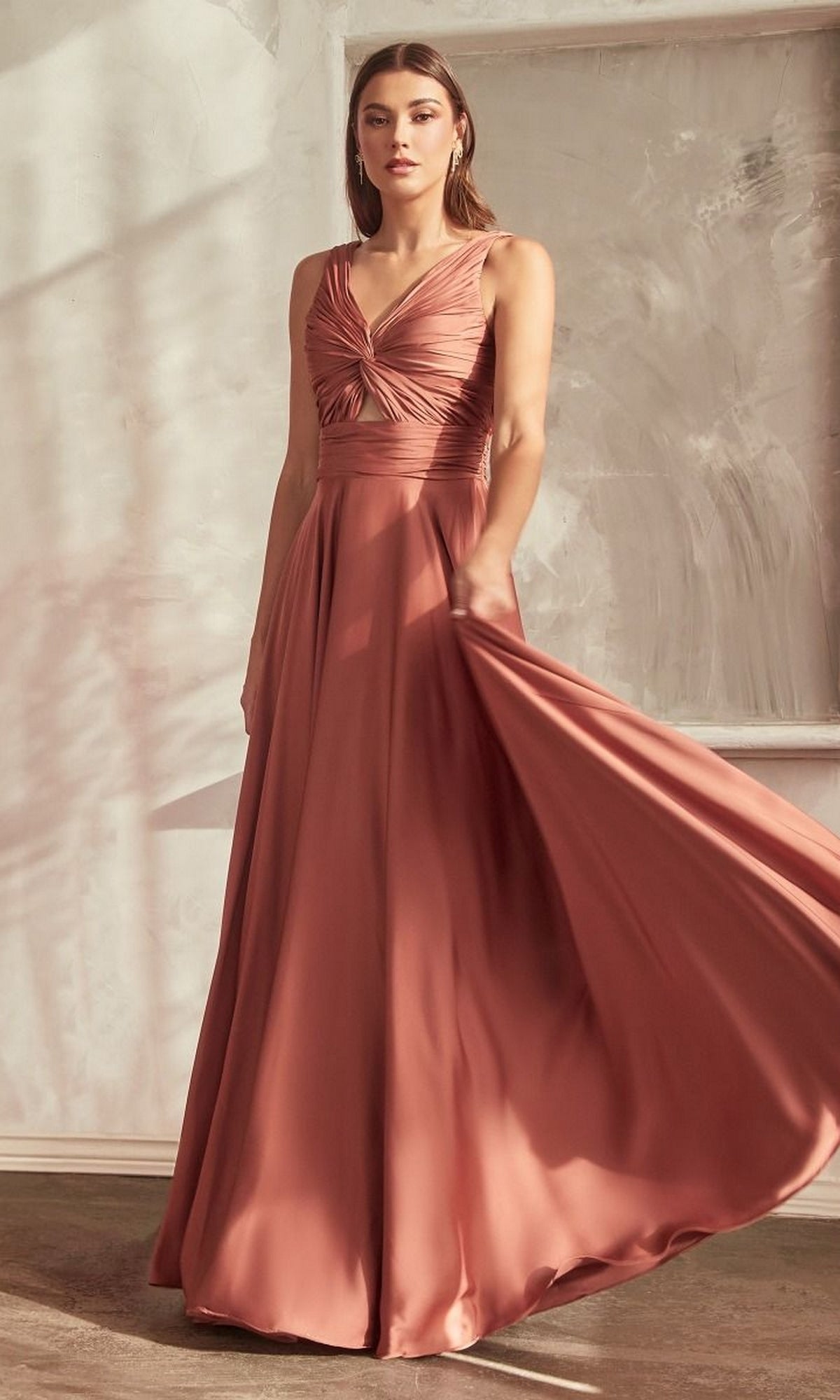 A woman stands indoors against a textured wall wearing the Twist-Front Long A-Line Cut-Out Prom Dress 7497—an affordable, sleeveless, floor-length A-line gown in dusty rose with a fitted bodice and flowing skirt.