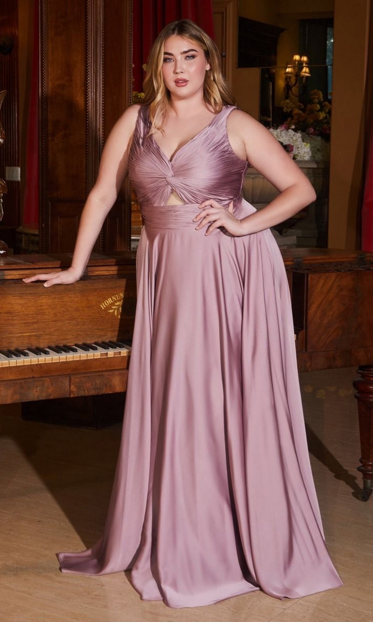 A woman in the Twist-Front Long Plus-Size Prom Dress 7497C, a sleeveless mauve gown, stands with one hand resting on a piano in an elegant room.