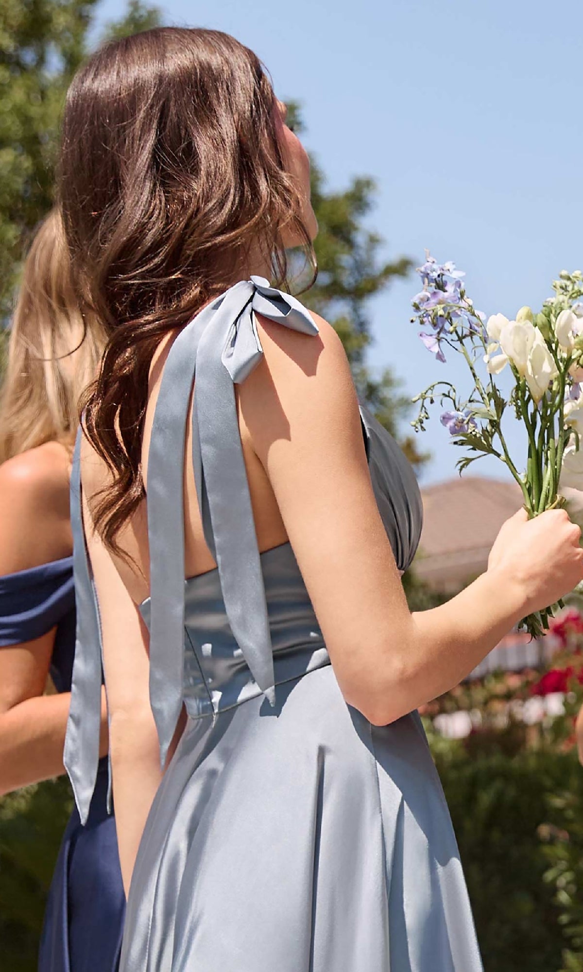 A woman in a light blue semi-formal dress with shoulder bows holds a bouquet outdoors; beside her stands another woman wearing the Ladivine 7499 tea-length wedding-guest dress in navy.