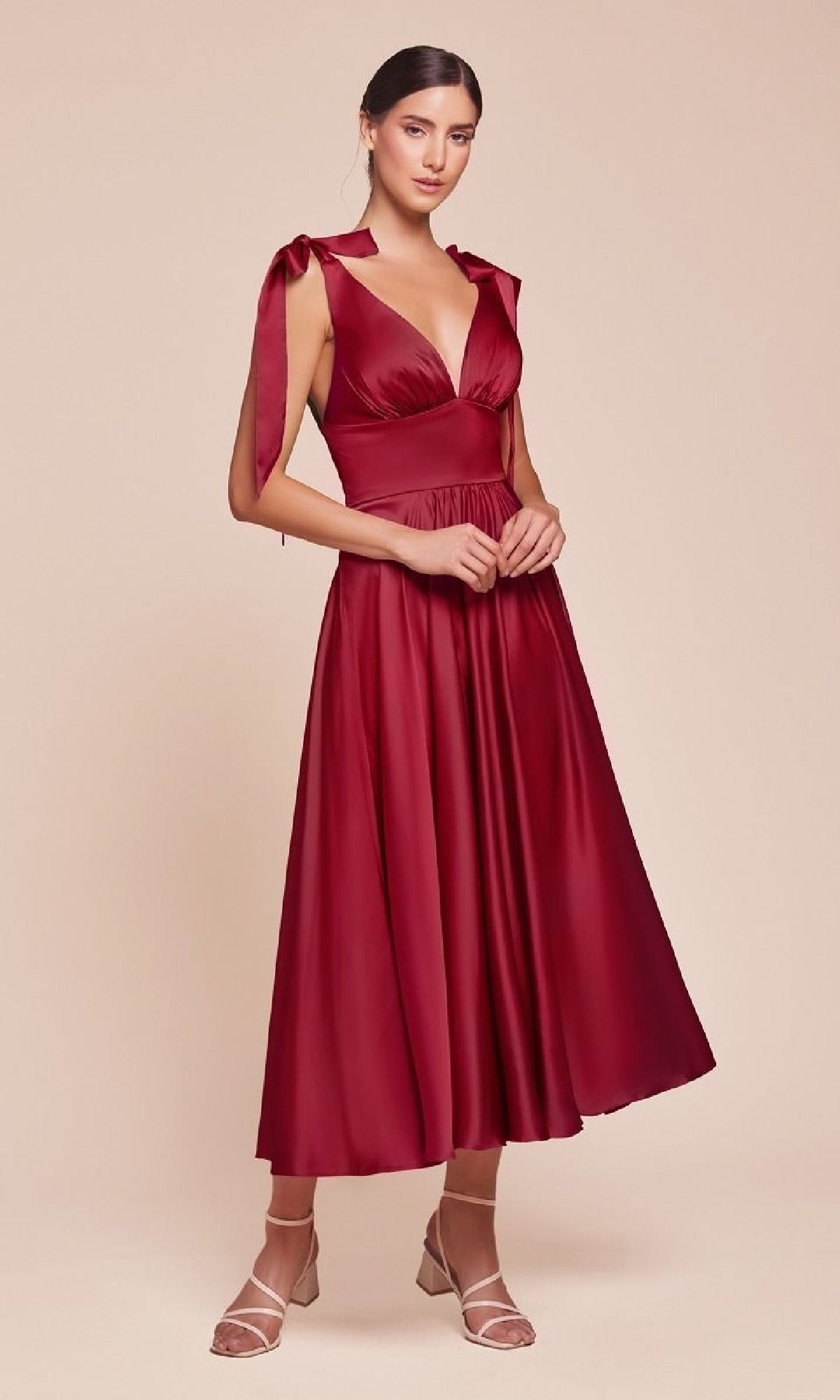 A woman models the Ladivine 7499 tea-length wedding-guest dress in deep red satin with tie straps and a V-neckline, paired with strappy nude heels, standing against a neutral background.