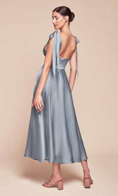Woman in the Ladivine 7499 Tea-Length Wedding-Guest Dress—light blue, backless satin A-line with tied shoulder straps—paired with nude heeled sandals, stands against a beige backdrop: a perfect semi-formal look for any occasion.