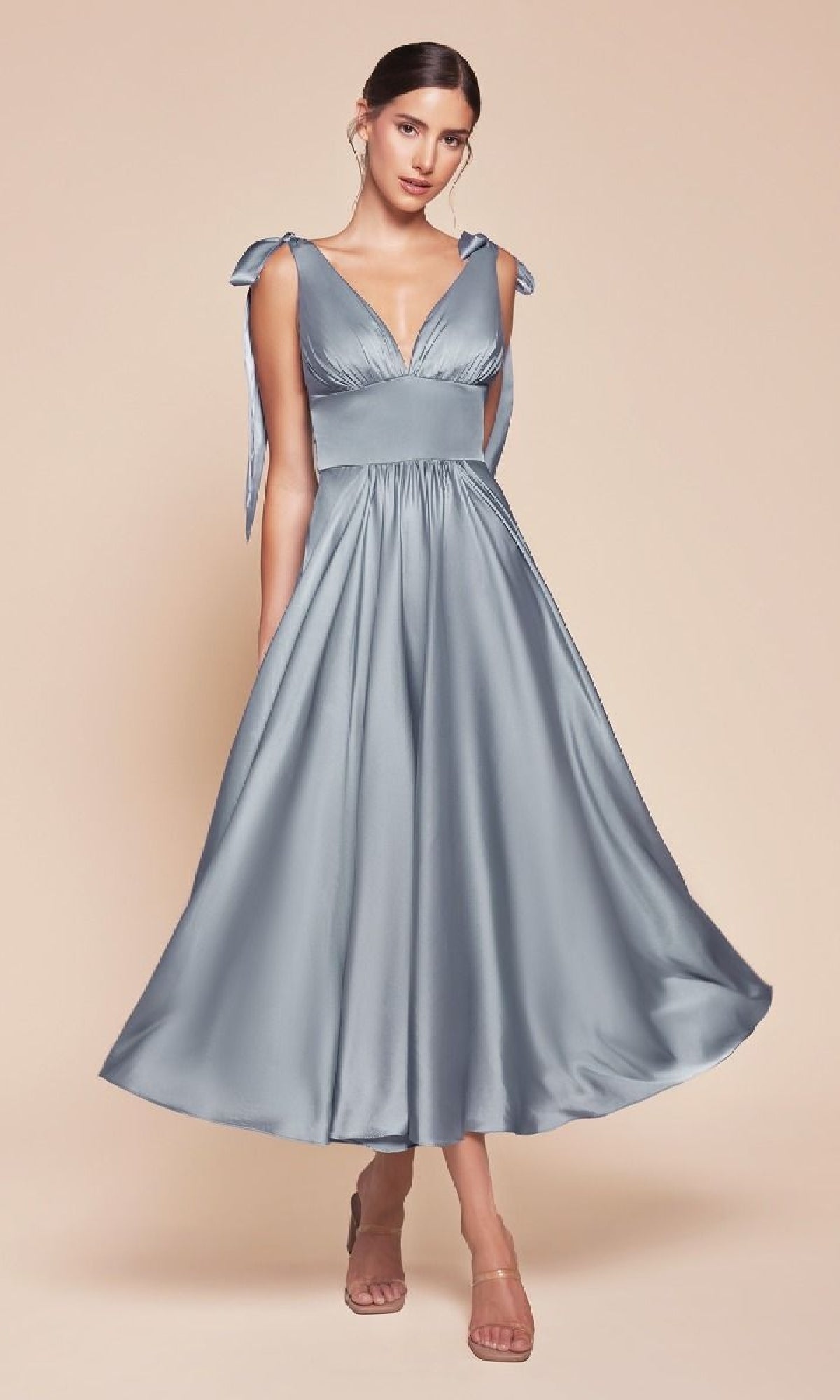 A woman wearing the Ladivine 7499 Tea-Length Wedding-Guest Dress—a light blue, sleeveless satin midi dress with a deep V-neck, tied shoulder straps, and flared skirt—stands against a plain beige background.