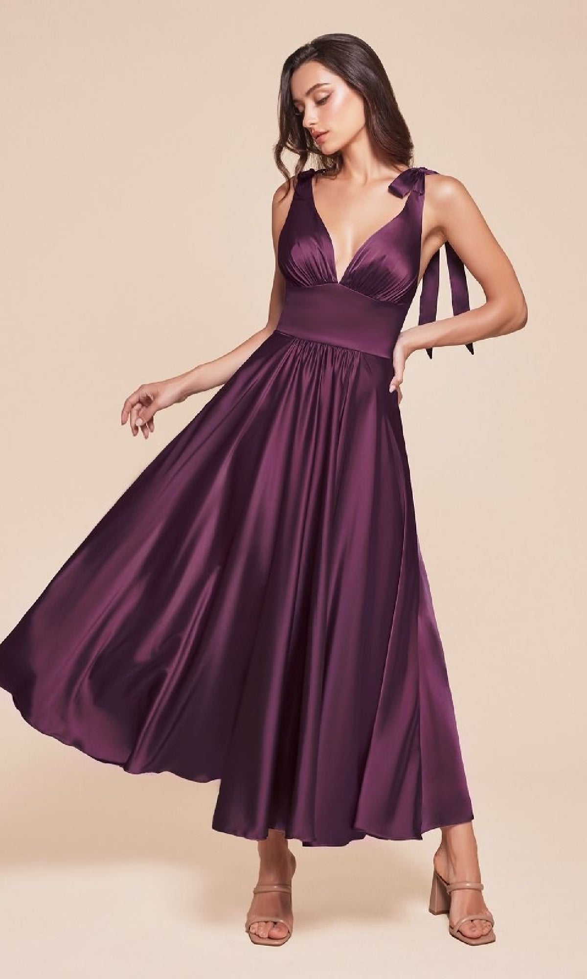A woman poses against a beige background in the Ladivine 7499 tea-length wedding-guest dress, featuring a sleeveless deep purple satin A-line silhouette with a V-neckline and bow shoulder straps.