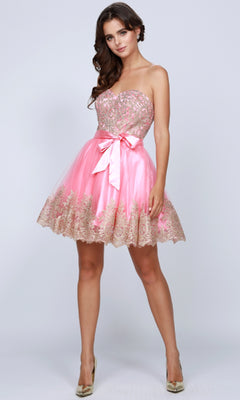 A woman stands against a light background in the Short Hoco Dress: Juliet 759—pink, strapless, with sequin and lace details, an A-line skirt, satin waist bow, and gold high heels.