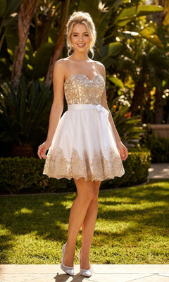 A woman poses outdoors in the Juliet 759 short hoco dress in white/gold, featuring a strapless gold embroidered bodice, white fit-and-flare skirt, and ribbon belt. She completes the look with matching heels against a green backdrop.