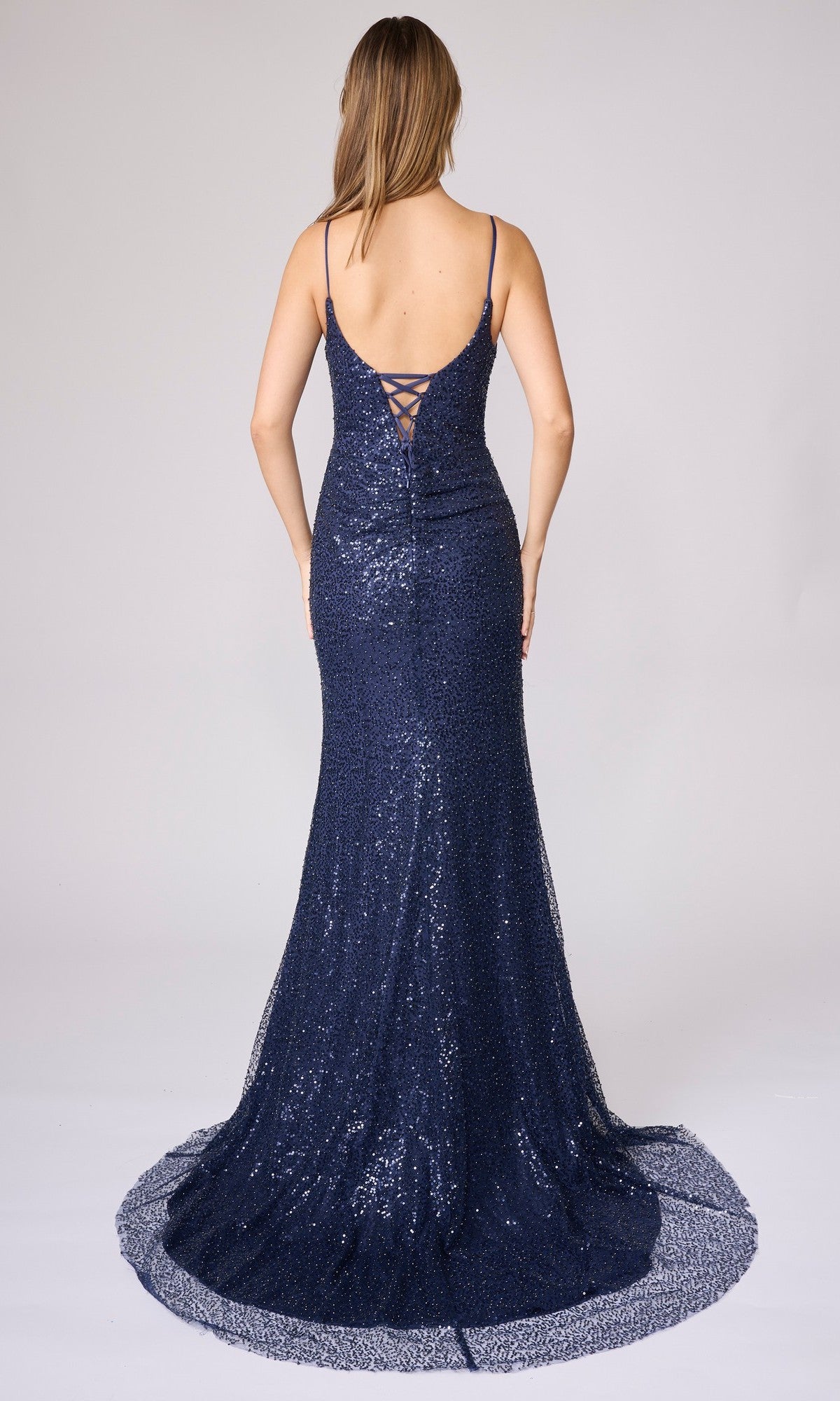 Woman in a navy blue sequin Nina Canacci 7656 long formal dress with spaghetti straps and lace-up back shimmers in this form-fitting look as she stands facing away from the camera.