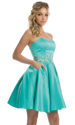 A woman poses with one hand in her pocket wearing the Juliet 769 short hoco dress—a turquoise, strapless homecoming dress with a sweetheart neckline, plus embroidery and beading detail at the waist—against a white background.
