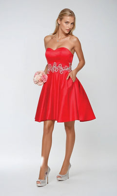A woman in the Short Hoco Dress: Juliet 769—strapless with a sweetheart neckline and floral belt detail—stands in silver high heels, holding a small bouquet of roses against a plain background.