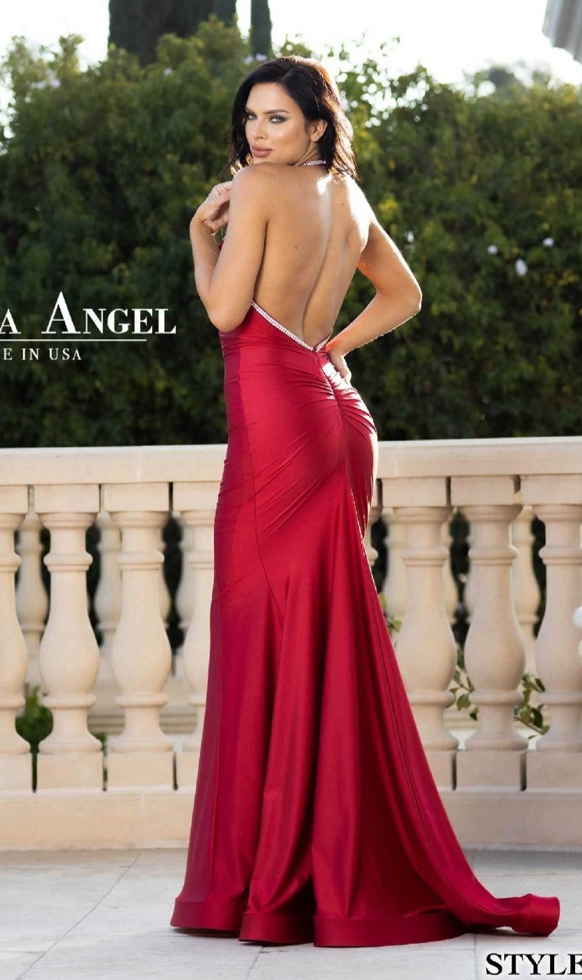 A woman stands on a patio in the Backless Long Halter Prom Dress: Jessica Angel 774, with a mermaid skirt, looking over her shoulder toward the camera. Shrubs and a balustrade are visible in the background.
