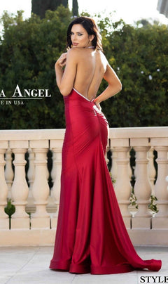 A woman stands on a patio in the Backless Long Halter Prom Dress: Jessica Angel 774, with a mermaid skirt, looking over her shoulder toward the camera. Shrubs and a balustrade are visible in the background.