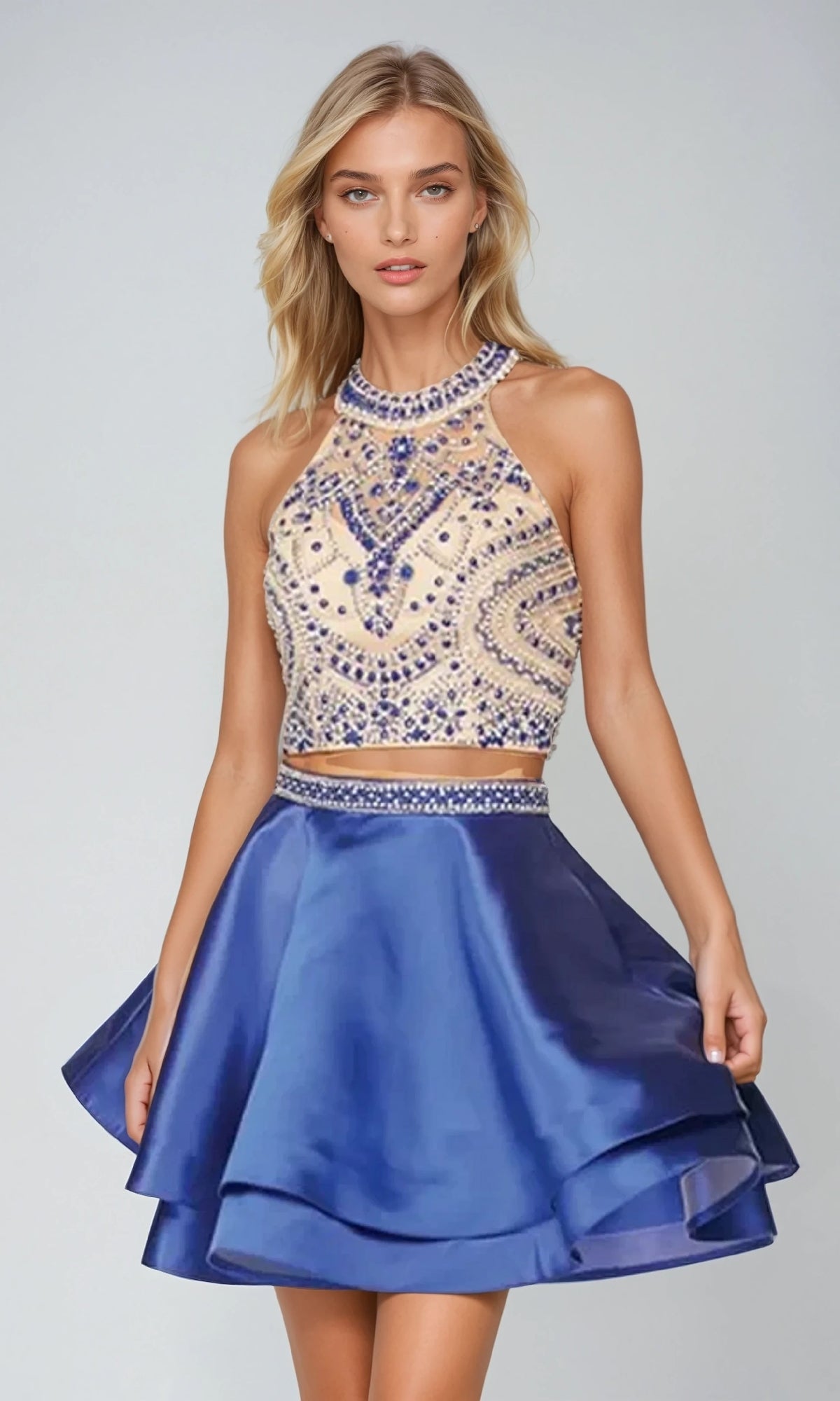 A woman models the Juliet 777 short party dress, featuring a sleeveless beaded top with intricate patterns and a shiny royal blue flared skirt, posing elegantly against a plain background.