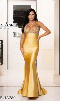 A woman stands indoors by a white door, wearing the Jessica Angel 780 Sheer-Neck Long Prom Dress in gold, featuring a fitted bodice and embellished straps.