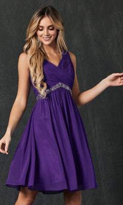 A person wearing the Juliet 786 short semi-formal dress—sleeveless, knee-length, with a pleated bodice and beaded waistband—stands against a dark background.