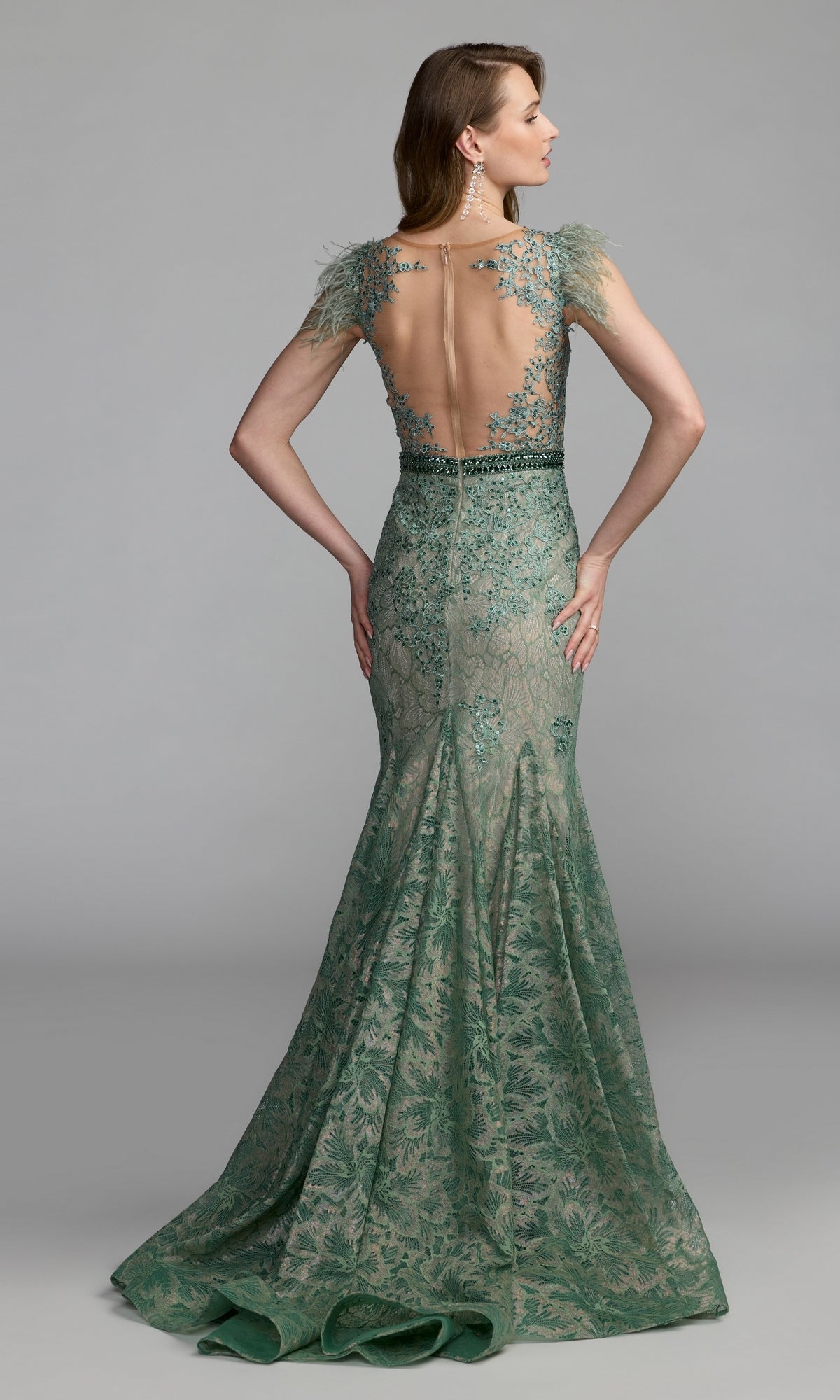 A woman wearing the Feathered Long Lace Formal Dress (Mac Duggal 79230), a fitted, floor-length green gown featuring lace detailing, a sheer back panel, and cap sleeves, stands against a plain gray background.