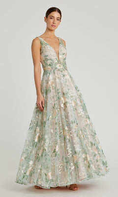 Woman models the Mac Duggal 79329 Long Cream Floral Prom Dress, a sleeveless, floor-length gown with a deep V-neckline and pastel floral embroidery, standing against a plain background.