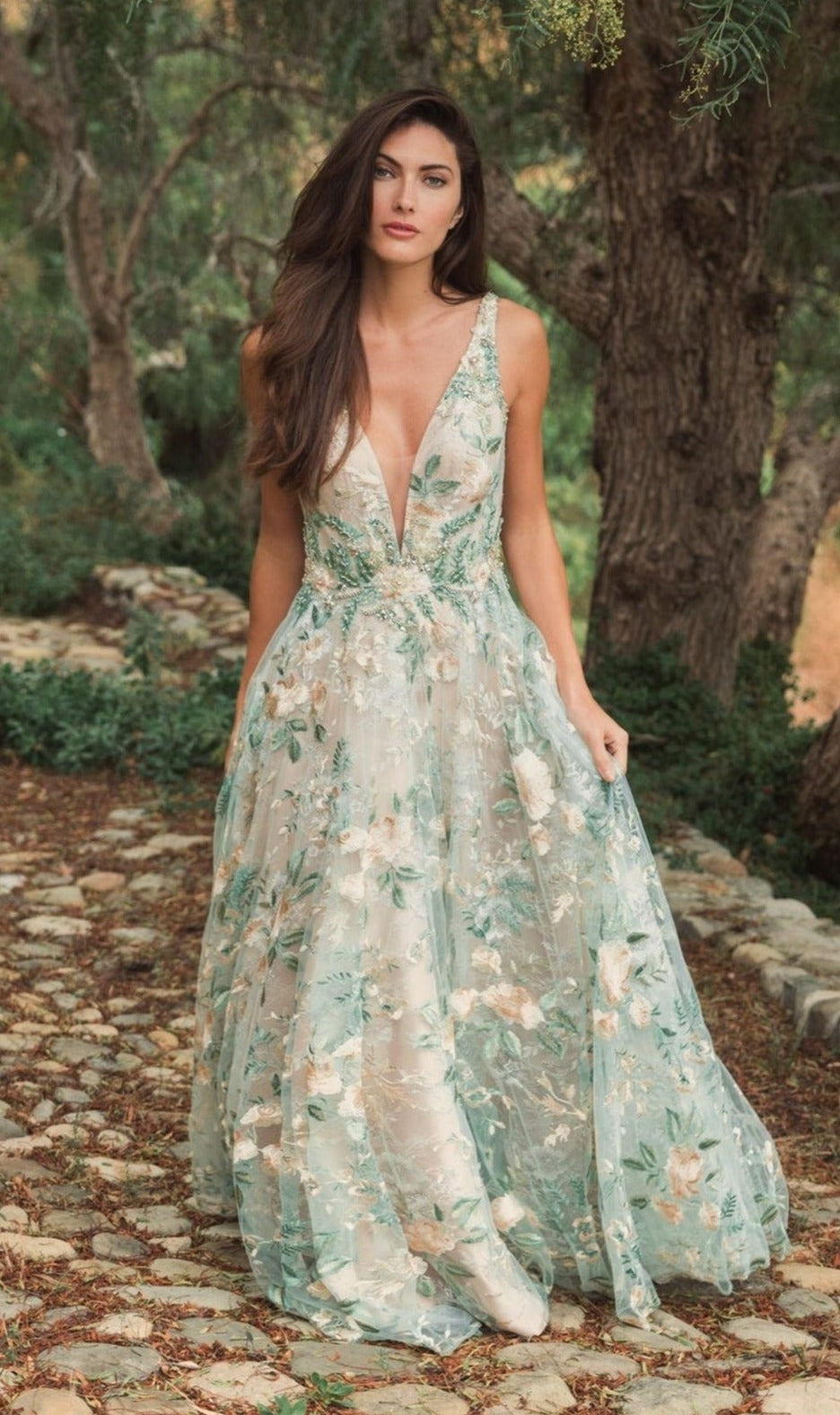 A woman stands on a stone path in a forest, wearing the Mac Duggal 79329—a long cream floral prom dress with embroidery and a deep V-neckline.