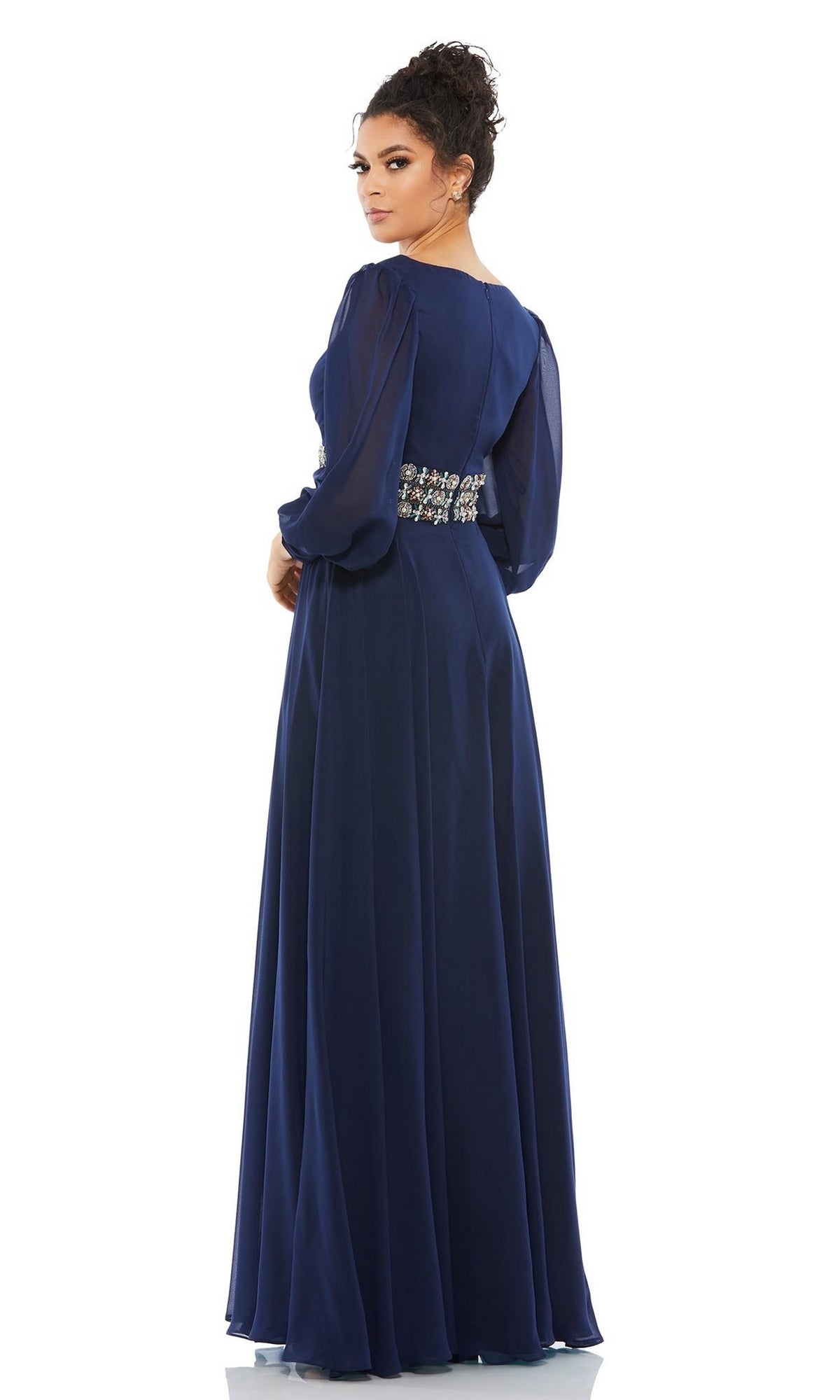 A woman in the Mac Duggal 79390 long formal dress, featuring navy blue fabric, sheer long sleeves, and an embellished waistband, stands gracefully looking over her shoulder.