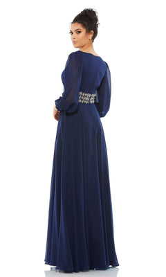 A woman in the Mac Duggal 79390 long formal dress, featuring navy blue fabric, sheer long sleeves, and an embellished waistband, stands gracefully looking over her shoulder.