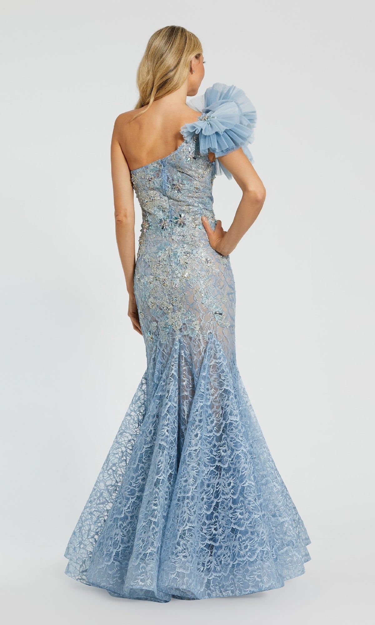A woman faces away, wearing the Mac Duggal 79456 Long Formal Dress—light blue lace with floral embellishments, a dramatic tulle one-shoulder detail, and an elegant mermaid silhouette.