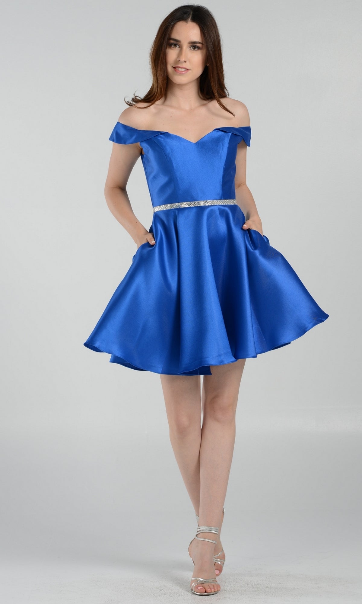 A woman in the Off-Shoulder Short Mikado Homecoming Dress 7948, a royal blue satin mini dress with a silver belt and silver strappy heels, stands against a light plain background.