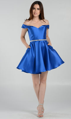 A woman in the Off-Shoulder Short Mikado Homecoming Dress 7948, a royal blue satin mini dress with a silver belt and silver strappy heels, stands against a light plain background.