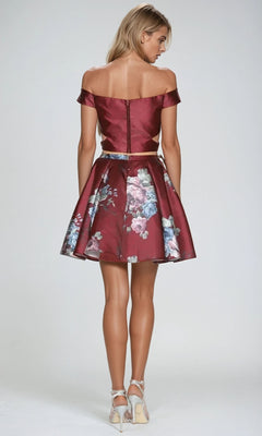 A woman faces away, wearing the Juliet 798 short party dress—an off-the-shoulder burgundy floral two-piece with an a-line skirt—paired with silver heels, set against a plain light background.