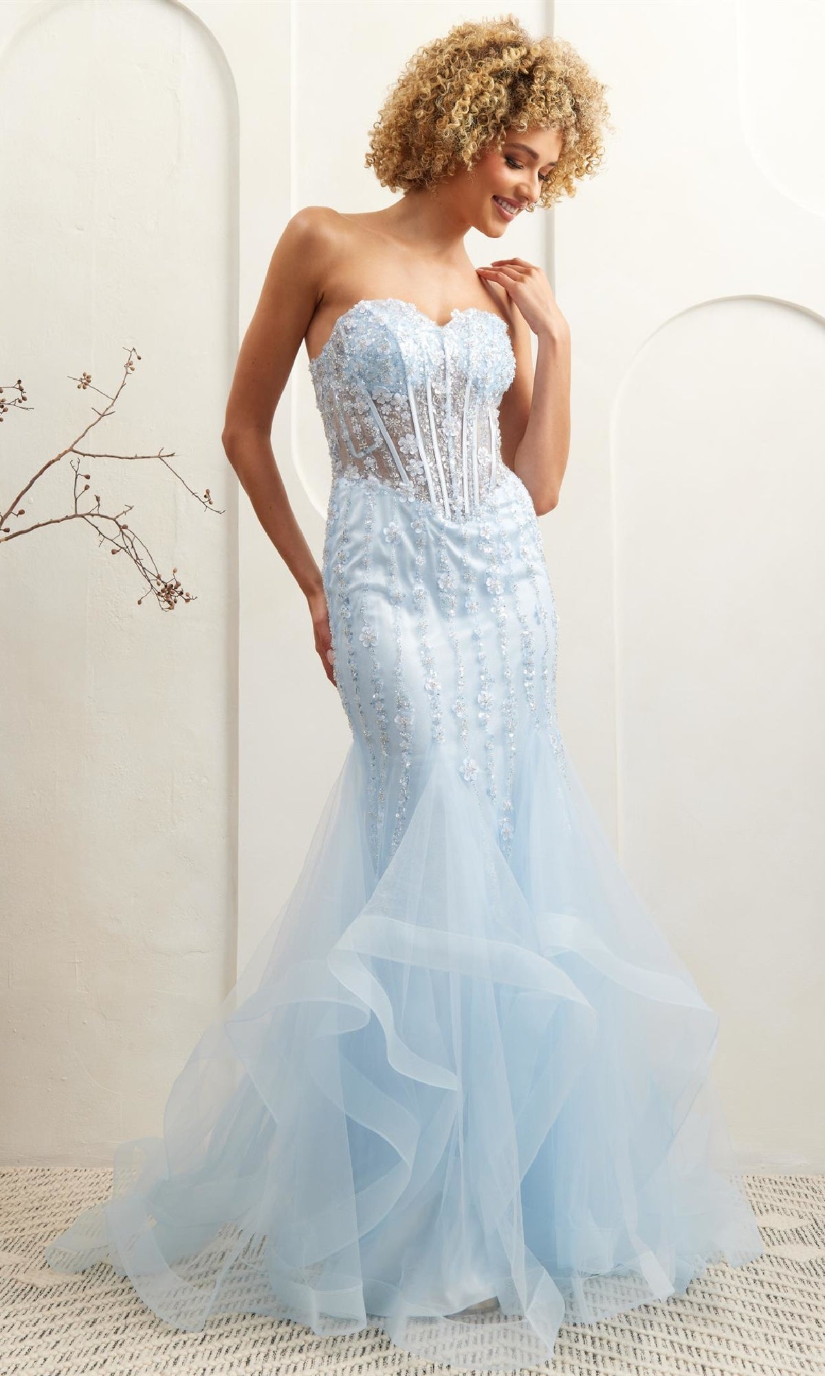 A woman wearing the Amelia Couture 8002 Strapless Mermaid Prom Dress in light blue with an embellished corset bodice and floral accents stands indoors, looking down and smiling.